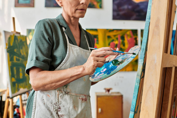 częściowy widok dojrzałej kobiety w fartuchu z paletą i malowanie pędzlem na sztaludze w pracowni artystycznej - Zdjęcie, obraz