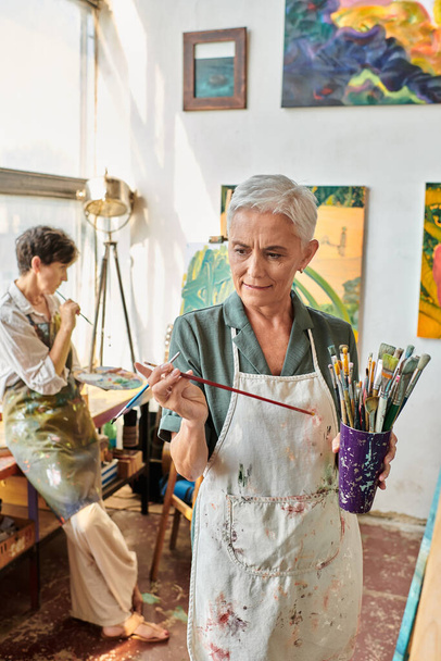 thoughtful mature woman choosing paintbrush near female friend in art workshop, creative hobby - Photo, Image