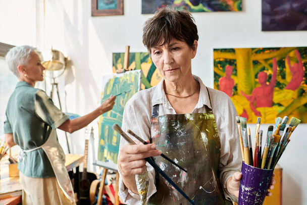 δημιουργική ώριμη γυναίκα ζωγράφος επιλέγοντας πινέλο κοντά θηλυκό φίλο ζωγραφική στο στούντιο τέχνης - Φωτογραφία, εικόνα