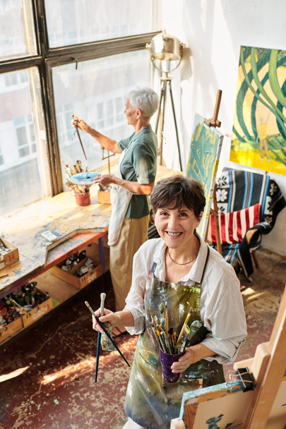 feliz mujer madura sosteniendo pinceles y mirando a la cámara cerca de una amiga en el taller de artesanía - Foto, Imagen
