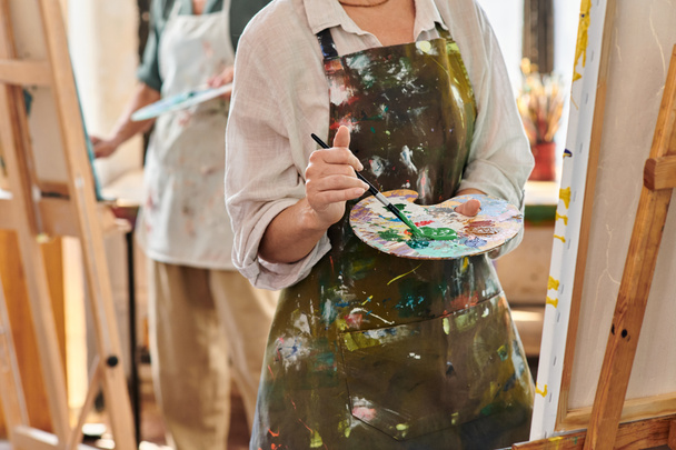 Ausgeschnittene Ansicht einer reifen Frau in Schürze, die Farben auf Farbpalette mischt, Meisterkurs in der Kunstwerkstatt - Foto, Bild