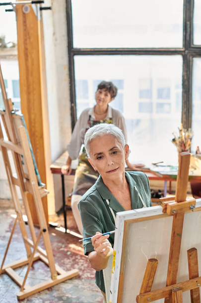 εμπνευσμένη ώριμη γυναίκα στην ποδιά ζωγραφική σε καβαλέτο στο master class στο εργαστήριο τέχνης, δημιουργικό χόμπι - Φωτογραφία, εικόνα