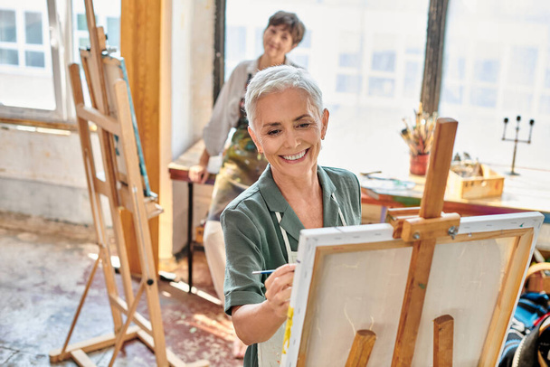 fröhliche reife Frau malt auf Staffelei neben qualifizierter Künstlerin in Werkstatt, Meisterkurs - Foto, Bild