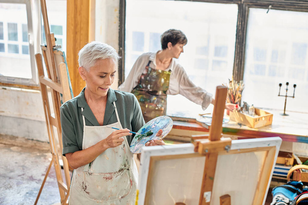 Lächeln reife Frau mischt Farben auf Farbpalette in der Nähe erfahrener Künstler in Werkstatt, Meisterkurs - Foto, Bild