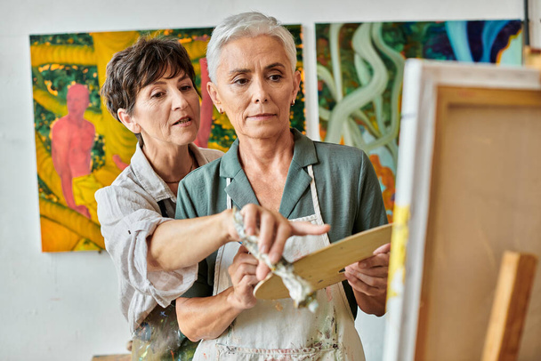 досвідчена художниця вказує на мольберт біля зрілої жінки під час майстер-класу в художній майстерні - Фото, зображення