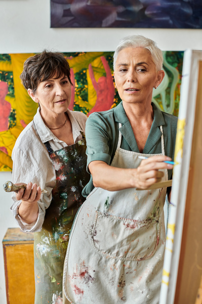 mature female artist pointing at easel near woman during master class in modern craft workshop - Photo, Image