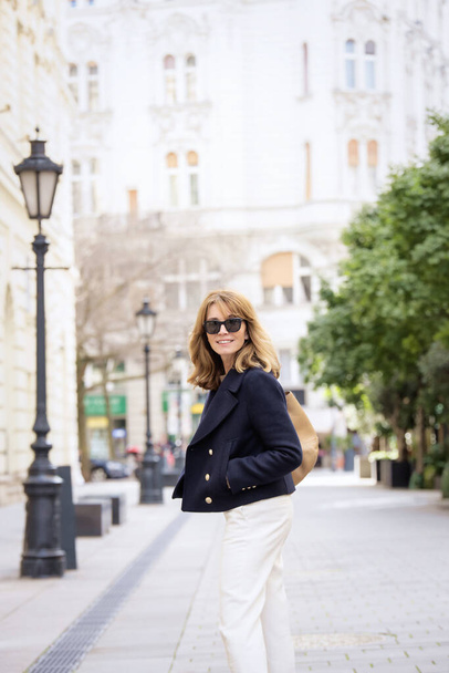 A middle-aged woman with blonde hair is walking down the street in the city centre. Attractive female wearing sunglasses and blue coat.  - Foto, Imagen