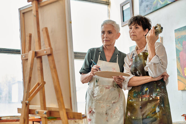 thoughtful and creative mature women looking at easel during painting master class in art workshop - Photo, Image