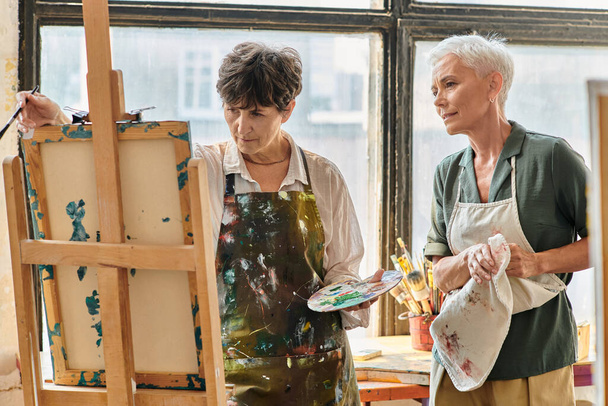 вдумлива зріла жінка дивиться на мольберт біля кваліфікованої жінки-художниці під час майстер-класу в студії - Фото, зображення