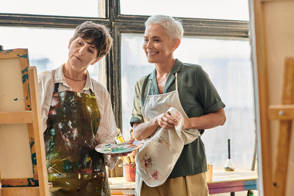 усміхнена зріла жінка дивиться на жінку-другу живопис на мольберті під час майстер-класу в студії - Фото, зображення