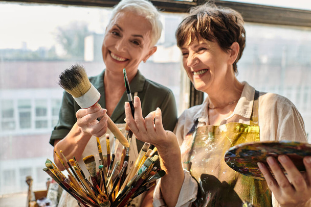 весела зріла жінка тримає різні пензлі під час майстер-класу в художній студії - Фото, зображення