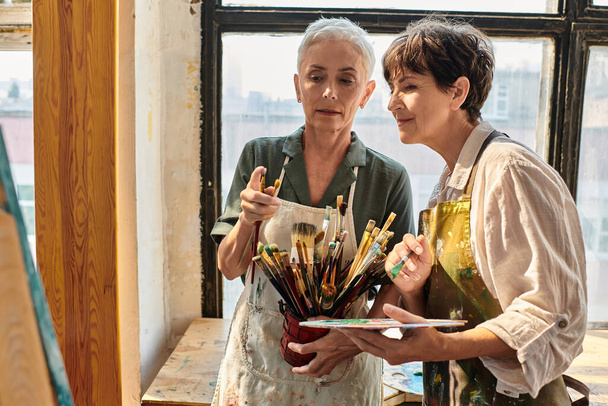 qualifizierte Künstlerin hilft reifen Frauen bei der Auswahl des Pinsels während eines Meisterkurses im Kunstatelier - Foto, Bild