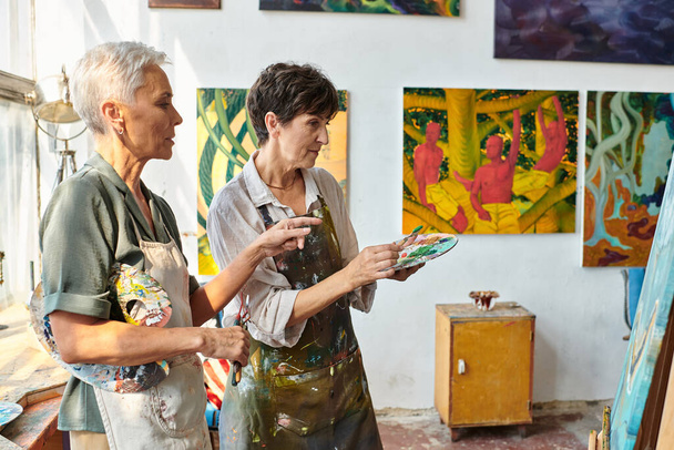 skilled female artist pointing at easel near mature woman with palette during master class in studio - Photo, Image