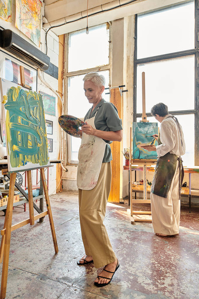 créative femme mûre en tablier peinture sur chevalet près d'amie dans moderne atelier d'art spacieux - Photo, image