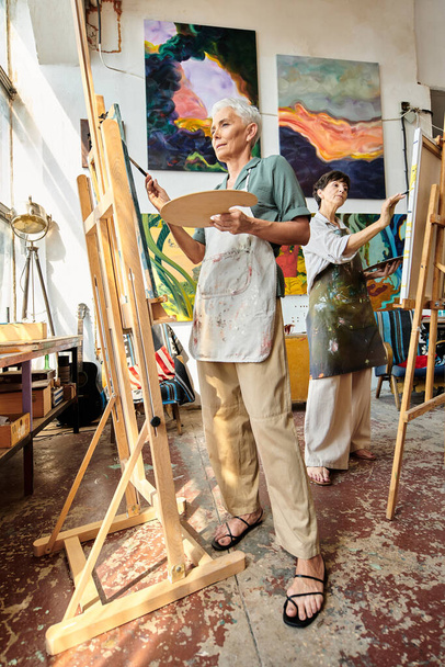 konzentrierte reife Frau in Schürzenmalerei auf Staffelei in moderner, geräumiger Werkstatt, künstlerisches Hobby - Foto, Bild