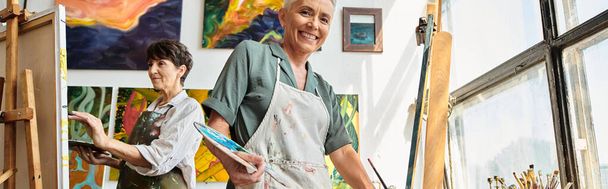 mulher madura feliz sorrindo para a câmera perto cavalete na oficina, hobby artístico, bandeira horizontal - Foto, Imagem