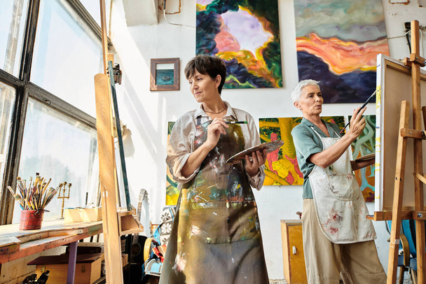 творческие зрелые художницы в фартуках, указывающие на мольберты в художественной студии, творческое хобби - Фото, изображение