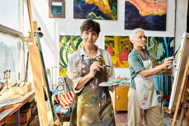 talented mature female artist with palette looking at camera near easel in studio, creative hobby - Photo, Image