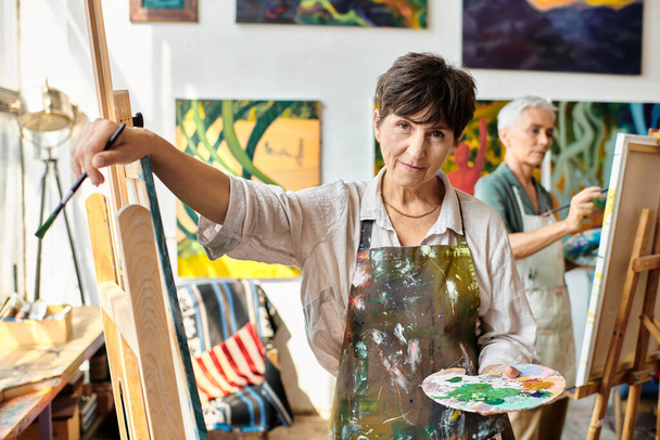 creative mature female artist with palette looking at camera near easel in studio, artistic hobby - Photo, Image
