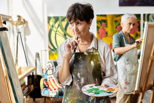 pensativo artista femenina madura con paleta mirando caballete en taller de arte, proceso creativo - Foto, Imagen