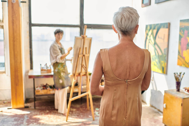 elegantní starší model pózující poblíž malby umělkyň na stojanu v umělecké dílně, tvůrčí proces - Fotografie, Obrázek