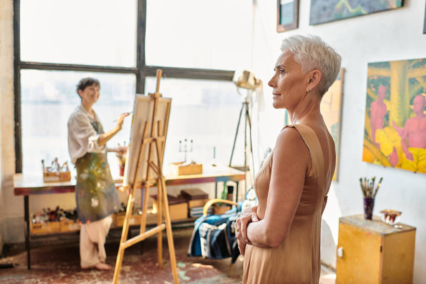 modelo maduro elegante posando cerca de artista femenina dolor en caballete en taller de arte, proceso creativo - Foto, Imagen