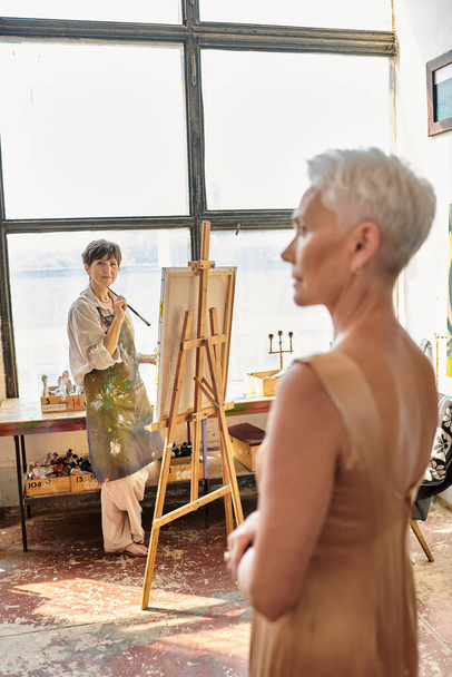 siro kypsä malli poseeraa lähellä naisartisti maalaus maalausteline työpajassa, taiteellinen prosessi - Valokuva, kuva