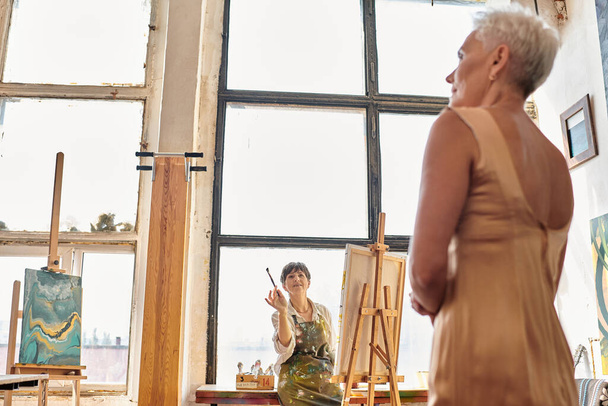 ammattimainen kypsä naistaiteilija puhuu mallille poseeraamalla taidetyöpajassa, luova prosessi - Valokuva, kuva