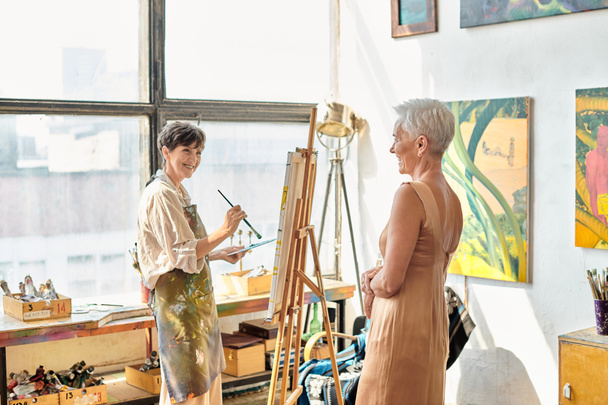 happy mature woman painting elegant female friend posing in art workshop, artistic process - Foto, Imagen