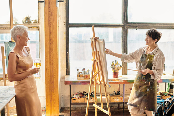 アートスタジオでワイングラスとポーズエレガントな女性の友人を描く幸せな成熟したアーティスト,創造性 - 写真・画像