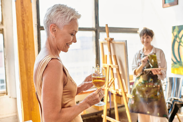 усміхнена модель середнього віку позує з келихом вина біля розмитої жінки-художниці в художній майстерні - Фото, зображення