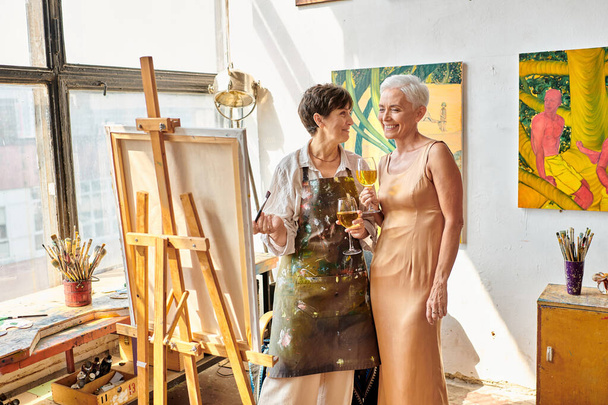 joyful female artist with elegant model standing with wine glasses near easel in art studio - Фото, изображение