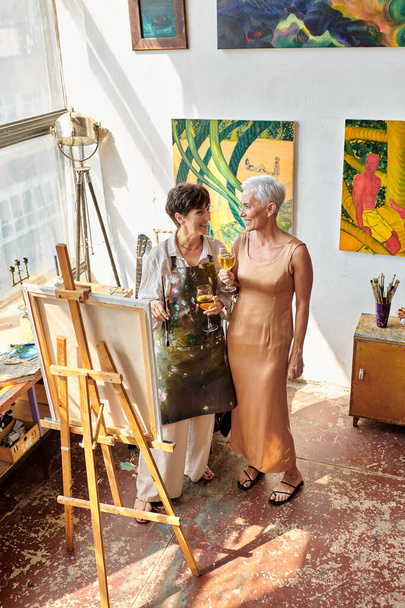 улыбающаяся женщина-художник со зрелой моделью, стоящая с бокалами вина в художественной студии, вид под высоким углом - Фото, изображение
