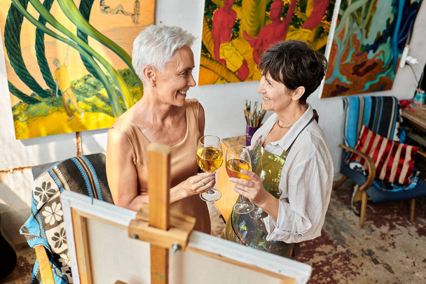 cheerful mature female friends with wine glasses near easel in art studio, artist and model teamwork - Photo, Image