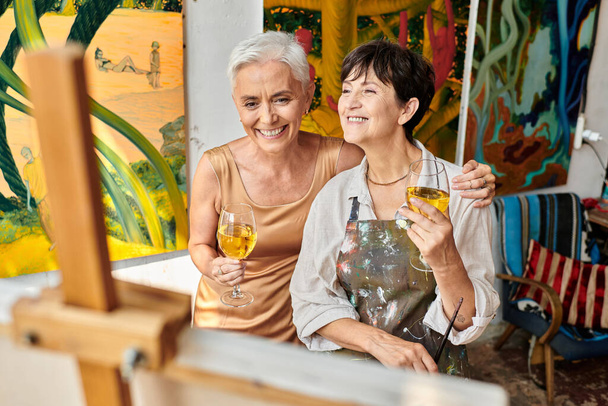 radosne dojrzałe kobiety z kieliszkami do wina uśmiechnięte w pobliżu sztalugi w warsztacie rzemieślniczym, artystka i modelka - Zdjęcie, obraz