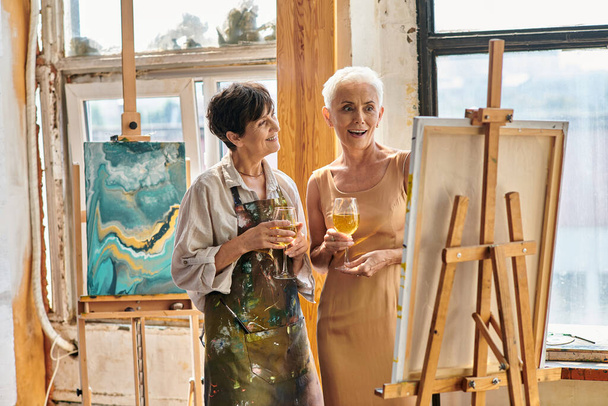 vaikuttunut kypsä malli viinilasin katselu maalausteline lähellä naisartisti työpajassa, taideteos - Valokuva, kuva