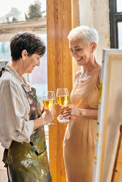 smiling female artist and elegant mature model clinking wine glasses in modern art workshop - Foto, Imagem