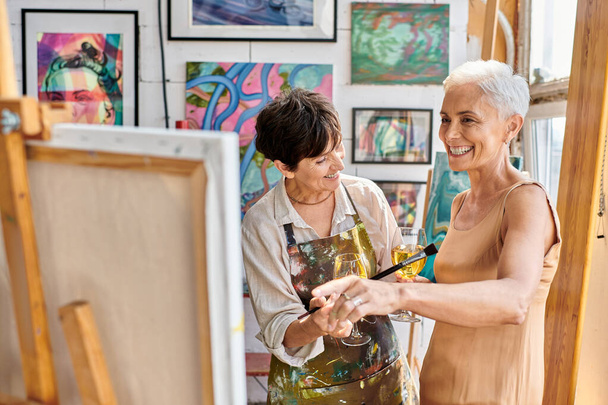 cheerful and stylish mature model with wine glass pointing at easel near woman painter in art studio - Photo, Image