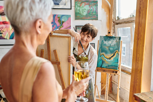 joyeuse femme mature artiste regardant flou modèle féminin posant avec verre à vin dans l'atelier - Photo, image