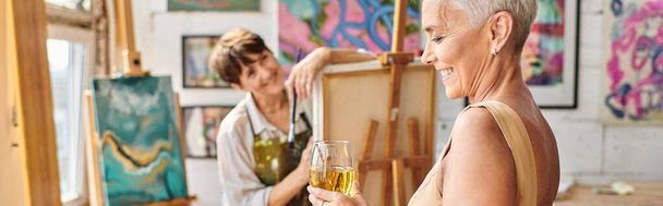 Şık, orta yaşlı, şarap kadehli bir kadın stüdyoda kadın sanatçının yanında gülümsüyor. - Fotoğraf, Görsel