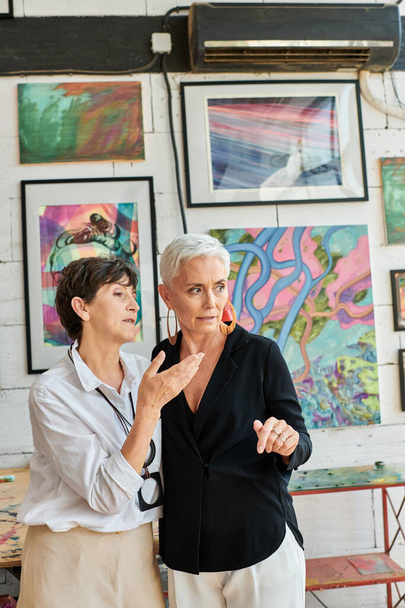 Kreatives gleichgeschlechtliches lesbisches Paar im Gespräch in der Nähe bunter Gemälde in der Werkstatt für moderne Kunst, Teamarbeit - Foto, Bild