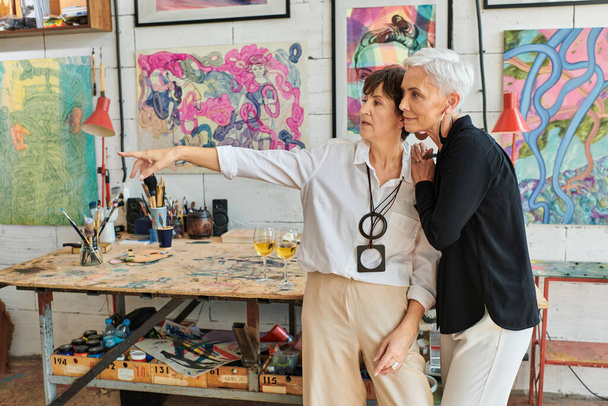 зрелая художница, указывающая рукой на картину и лесбиянку-партнера в мастерской современного ремесла - Фото, изображение