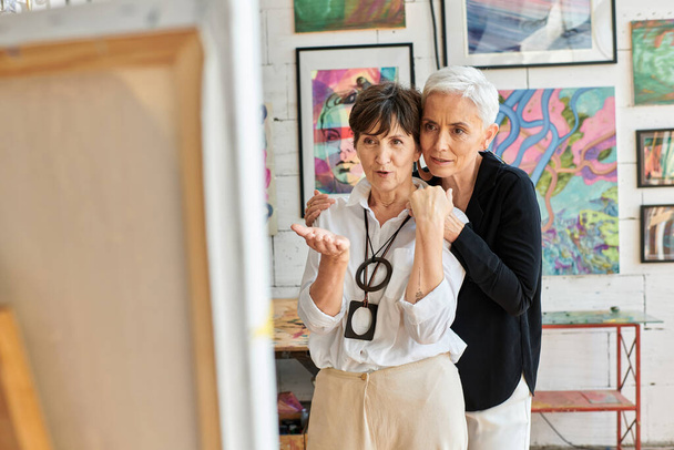 impressed middle aged female artist pointing with hand near lesbian partner in modern art studio - Photo, Image