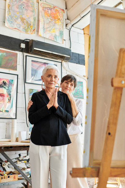 impressed female artist looking at easel near lesbian partner in workshop, teamwork and art - Photo, Image