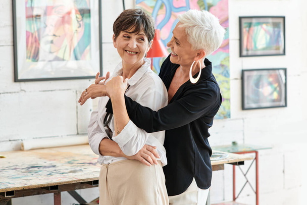 radosna i modna dojrzała kobieta obejmująca lesbijkę partnerkę w warsztacie sztuki nowoczesnej, szczęście - Zdjęcie, obraz