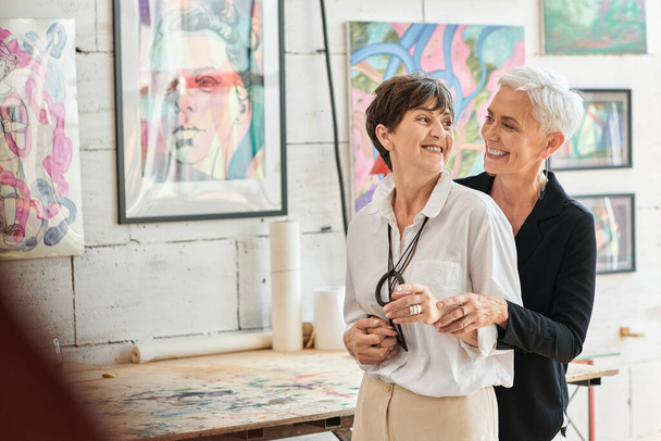 fröhliche lesbische Künstler, die einander in der Werkstatt für moderne Kunst umarmen und anschauen, - Foto, Bild