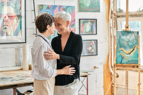 gelukkig en modieus lesbische kunstenaars omarmen in moderne kunst studio met creatieve schilderijen - Foto, afbeelding