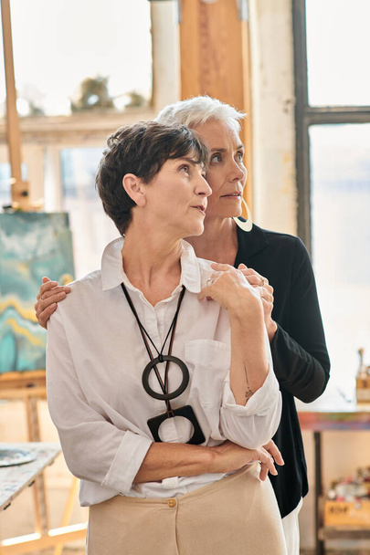 thoughtful and stylish lesbian artists looking away in modern art studio with creative paintings - Photo, Image