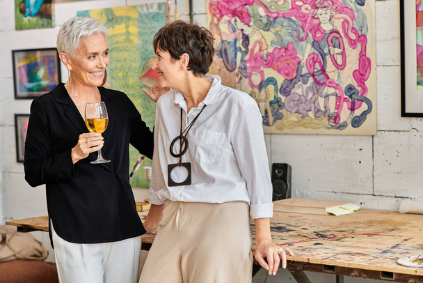 radostná a stylová zralá žena se sklenkou na víno mluví s lesbickým partnerem v moderním uměleckém studiu - Fotografie, Obrázek