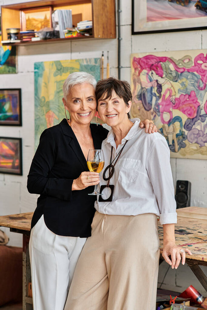 лесбийская пара художников с бокалом вина обнимает и смотрит в камеру в студии современного искусства - Фото, изображение